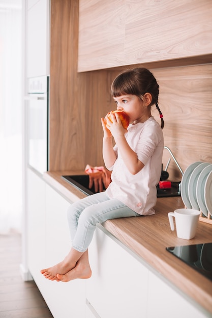 Schattig klein meisje bijten rode appel zittend op een keukenoppervlak. Gezond eten concept.