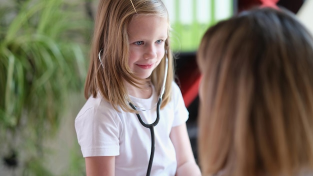 Schattig klein meisje controleert organen van vrouw met stethoscoop met glimlach op gezicht schattige dokter