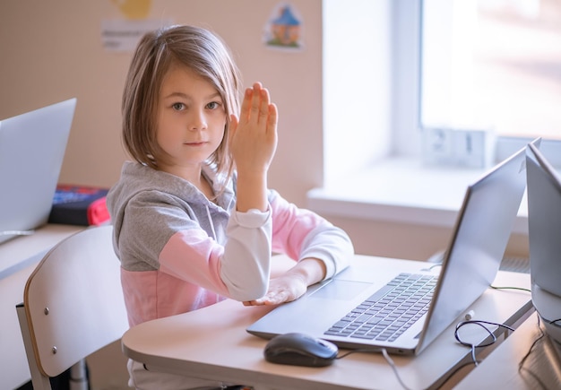 Schattig klein meisje dat aan de schoolbank zit met een laptop, steekt zijn hand op om te antwoorden