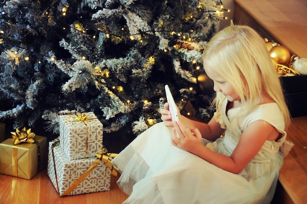 Schattig klein meisje dat digitale tablet gebruikt terwijl ze bij de kerstboom zit