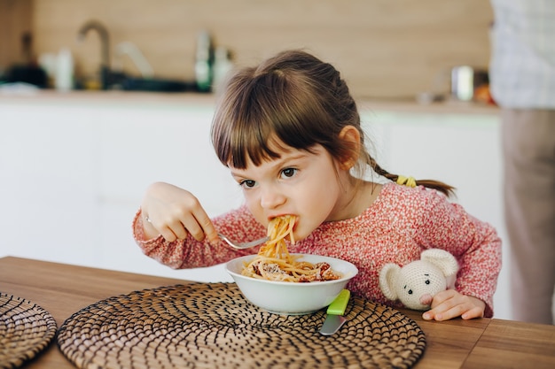 Schattig klein meisje dat spaghetti met saus eet, zittend in een keuken thuis