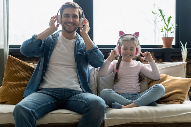 Schattig klein meisje en haar knappe vader in koptelefoon luisteren naar muziek, glimlachen, zittend op de bank thuis.