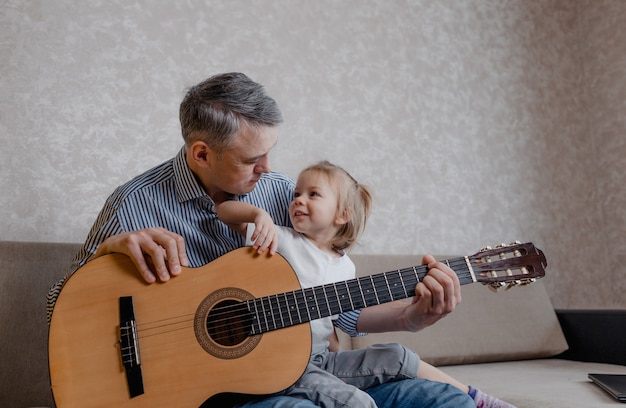 Schattig klein meisje en haar knappe vader spelen gitaar en glimlachen thuis op de bank. Vaderdag. zorg en opvoeding van kinderen.