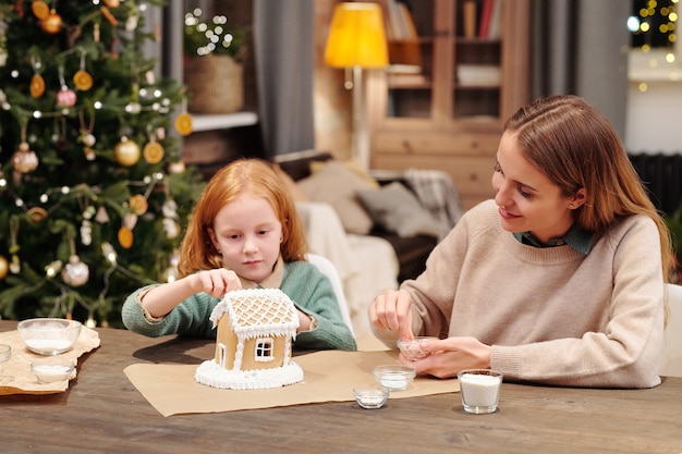 Schattig klein meisje en haar moeder beregening dak van peperkoek huis versierd met slagroom tijdens het bereiden van feestelijk dessert