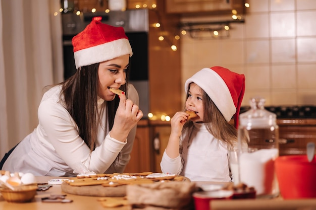 Schattig klein meisje en haar moeder in kerstmuts die zelfgemaakte peperkoek eet en gelukkig klein lacht