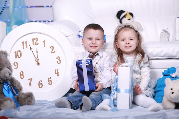 Foto schattig klein meisje en jongen met geschenkdozen en grote witte klok zitten in de buurt van de witte bank in kerstmis en nieuwjaar