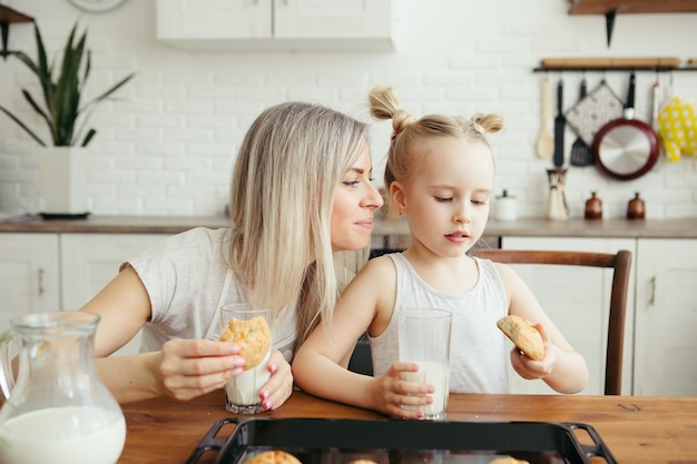 Schattig klein meisje en moeder die versgebakken koekjes met melk eten in de keuken. Gelukkig gezin. Tonen.