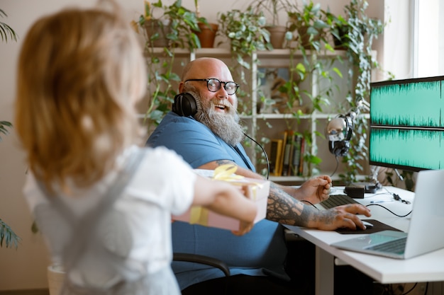 Schattig klein meisje geeft cadeau aan verraste vader die op de computer in een lichte kamer werkt