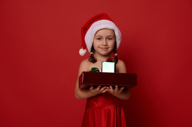 Schattig klein meisje gekleed in santa carnaval kostuum, met geschenken op haar handen, glimlacht camera kijken, poseren tegen rode achtergrond. vrolijk kerst- en nieuwjaarsconcept met kopieerruimte voor advertentie