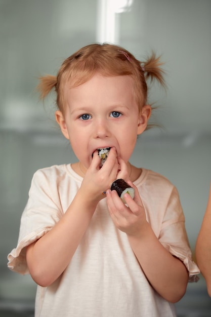 Schattig klein meisje geniet van het eten van sushi Vrolijk, grappig kaukasisch blond peutermeisje met sushibroodje en op keukenachtergrond