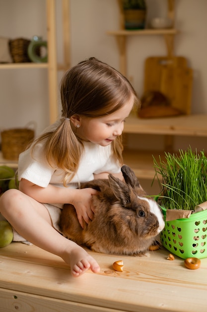 Schattig klein meisje in de houten keuken van het huis voedt het konijn vers gras