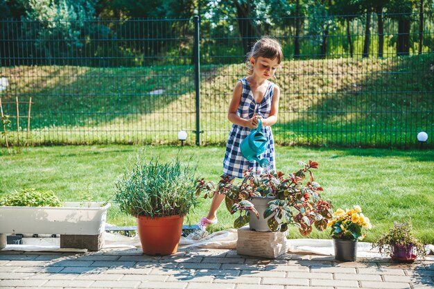 Schattig klein meisje in een blauwe geruite jurk die de planten water geeft uit een gieter in de tuin. Outdoor activiteit kinderen.