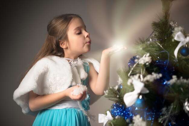 Schattig klein meisje in een blauwe jurk heeft een geschenk in haar handen bij een kerstboom