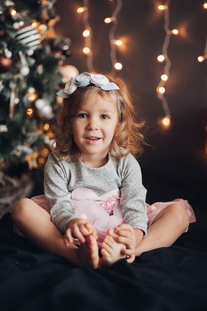 Schattig klein meisje in een elegante jurk camera met kerstboom en verlichting kijken