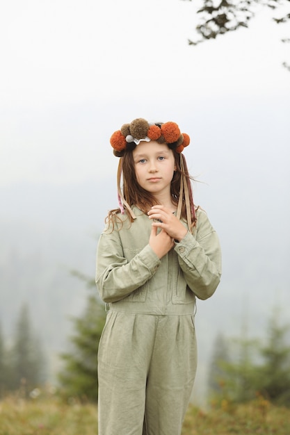 Foto schattig klein meisje in een pastel jumpsuit met een kleurrijke krans op haar hoofd. portret van een klein meisje buiten in de mistige bergen