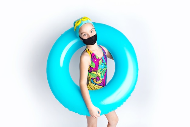 Schattig klein meisje in een strandzwempak en een opblaasbare turquoise zwemcirkel, in een zwart beschermend covid-19-masker. Recreatieconcept tijdens het coronavirus. Hoge kwaliteit foto
