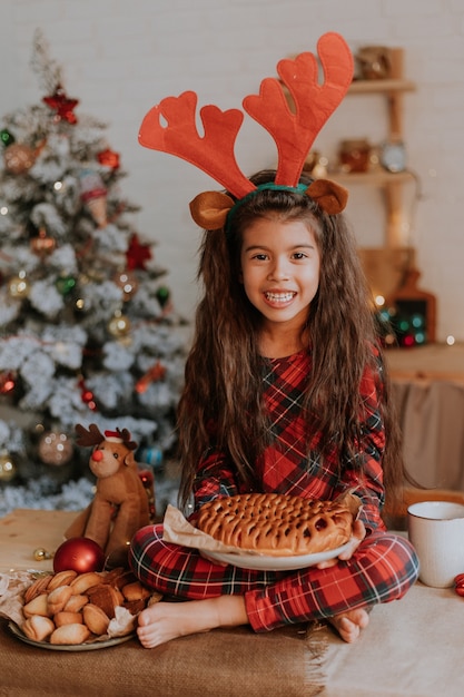 Schattig klein meisje in geruite rode pyjama's met hertenhoorns op haar hoofd eet een kersttaart