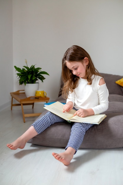 schattig klein meisje in jeans en een witte coltrui leest en doet huiswerk zittend op het bed