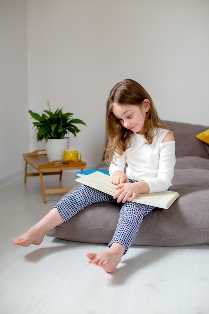 schattig klein meisje in jeans en een witte coltrui leest en doet huiswerk zittend op het bed