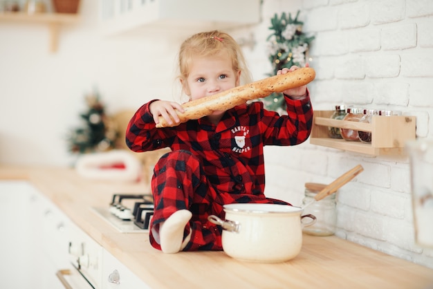 Schattig klein meisje in kerstmuts, koekjes in de keuken thuis voorbereiden. zit op de keukentafel en helpt moeder met het bereiden van een feestelijk kerstdiner