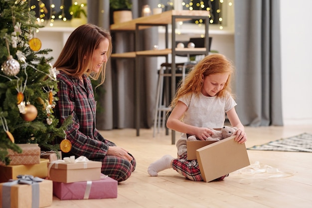 Schattig klein meisje in pyjama die teddybeer uit de geschenkdoos haalt terwijl ze op eerste kerstdag op de vloer naast haar moeder zit door versierde spar