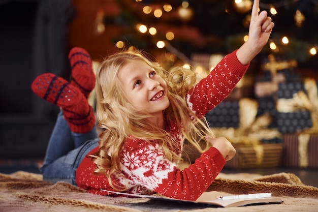 Schattig klein meisje in rode feestelijke trui binnenshuis liggen vieren nieuwjaar en kerstvakantie.