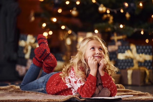 Schattig klein meisje in rode feestelijke trui liggend met teddybeer binnenshuis Nieuwjaar vieren.