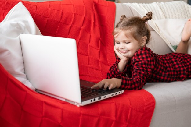 schattig klein meisje in rode kerstpyjama met laptop, videochat, liggend op de bank