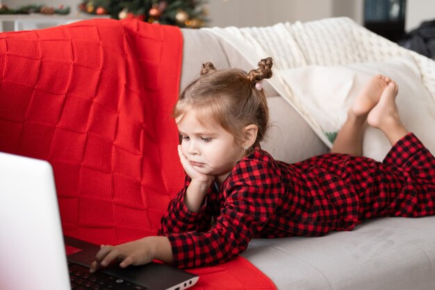 schattig klein meisje in rode kerstpyjama met laptop, videochat, liggend op de bank
