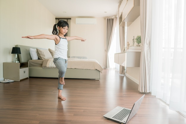 Schattig klein meisje in sportkleding die online video op laptop bekijkt en thuis fitnessoefeningen doet.