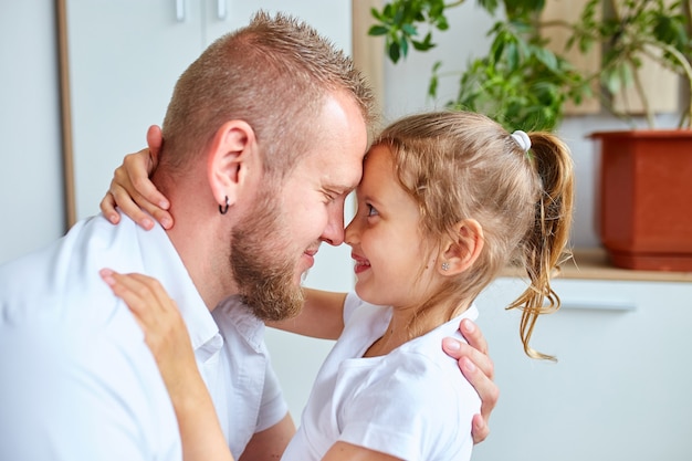 Schattig klein meisje in witte jurk knuffelen liefhebbende vader die naar hem kijkt met liefde en tederheid, vader en dochter brengen tijd thuis door.