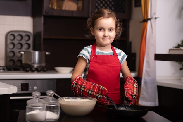 Schattig klein meisje kind in rode schort in de keuken thuis deeg maken voor pannenkoeken gebak kinderen koken concept