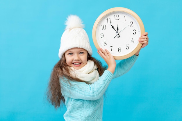 Schattig klein meisje kind in winterkleren muts en trui met een horloge in haar handen op een blauwe geïsoleerde achtergrond glimlachen, een plaats of ruimte voor tekst