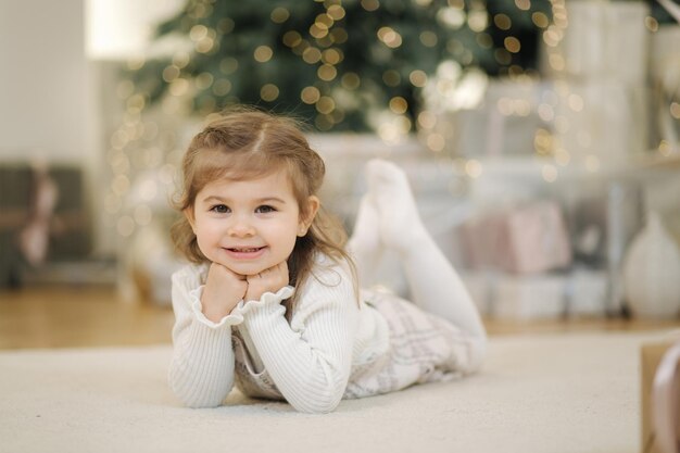 Schattig klein meisje liggend op haar voorkant in de buurt van dennenboom en lacht kerststemming thuis met veel