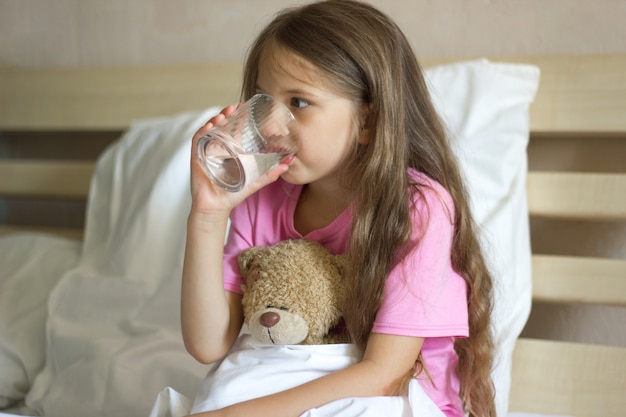 schattig klein meisje met blond zit op het bed, houdt een glas water vast, drinkt water en knuffelt teddybeer