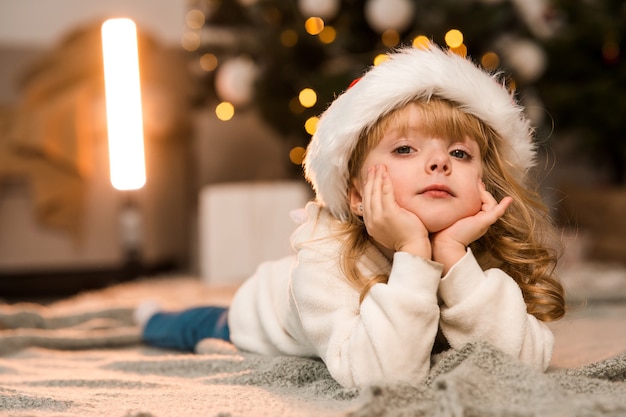 Schattig klein meisje met een kerstmuts ligt op de vloer in de kamer tegen de achtergrond van een kerstboom