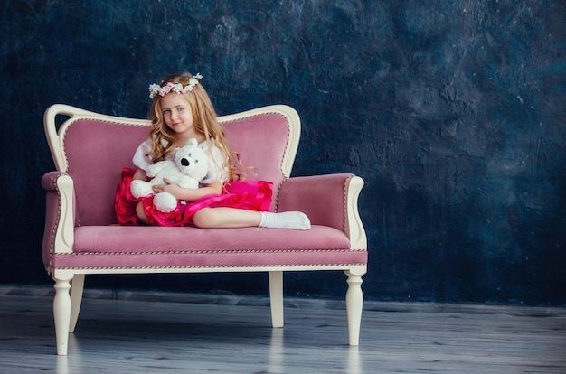 Schattig klein meisje met een krans op haar hoofd zittend op een roze bank met een witte teddybeer tegen de donkere muur.