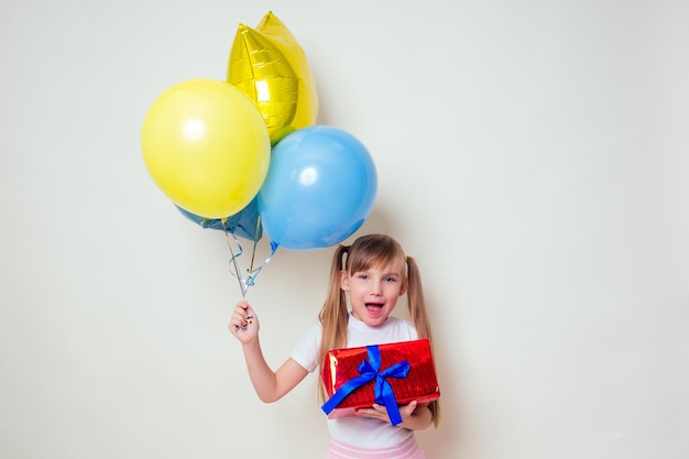 Schattig klein meisje met helium blauw gele en gouden ballonnen en een doos met een gelukkige verjaardag cadeau op witte achtergrond in studio Kerstmis en Nieuwjaar vakantie