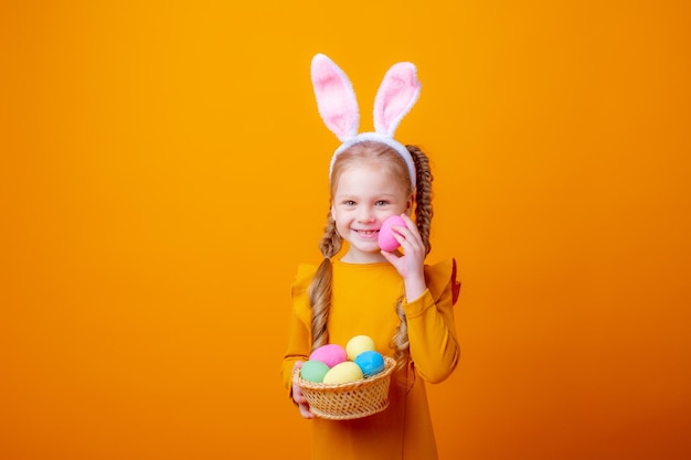 Schattig klein meisje met konijnenoren houdt een mand met paaseieren op een gele achtergrond