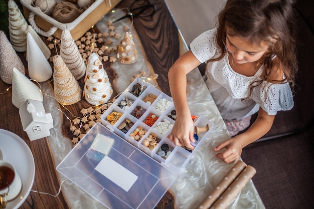 Schattig klein meisje met krullend haar knutselen en kerstboomkegel versieren met knopen