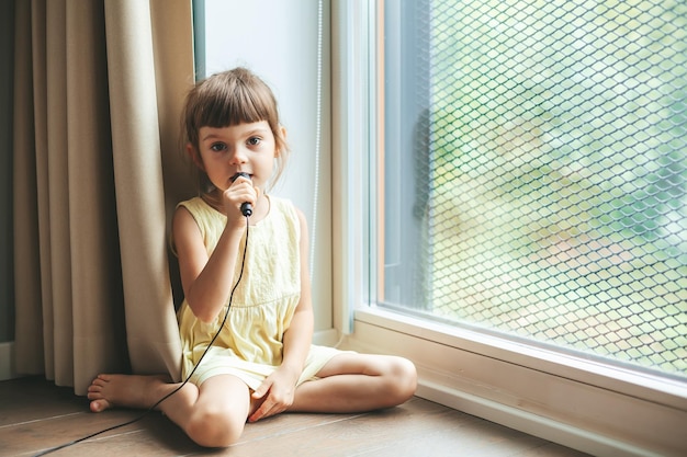 Schattig klein meisje met microfoon zingen thuis zittend op de vloer bij het raam