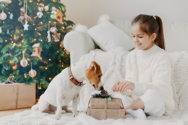 Schattig klein meisje met paardenstaart, gekleed in witte wintertrui, zit gekruiste benen op de vloer, pakt kerstcadeau uit