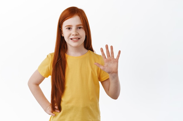 Schattig klein meisje met sproeten en rood gemberhaar dat hallo zegt. kind zwaaiend met de hand om hallo te zeggen, hey what's up gebaar, begroet vriend op speelplaats, witte achtergrond