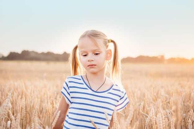 Schattig klein meisje met twee paardenstaarten in het gele tarweveld bij zonsondergang zomer landschap, zomer landbouw achtergrond met rijpe tarwe spikes