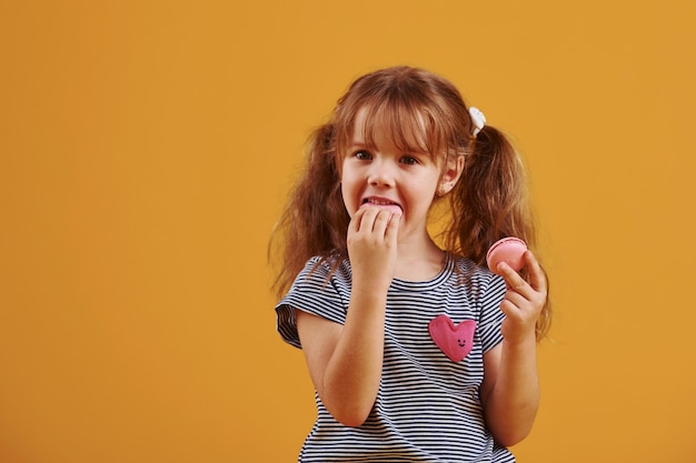 Schattig klein meisje met zoet eten in de studio tegen gele achtergrond.