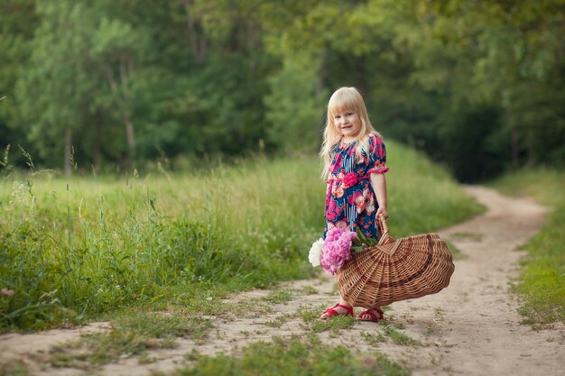 Schattig klein meisje op een bosweg met een mand met bloemen Het concept van zorgeloze jeugd