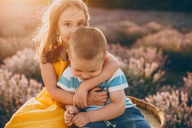 Schattig klein meisje terwijl ze haar broertje kust en hem omhelst tegen zonsondergang in een veld met bloemen.