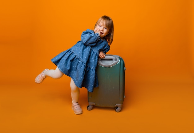 Schattig klein meisje van 4 jaar oud in een blauwe denim jurk met een groene koffer op een gele achtergrond.