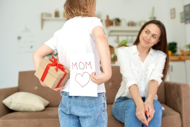 Schattig klein meisje verbergt een cadeau en een cadeaubon voor haar moeder achter haar rug close-up Moeder kijkt uit naar het cadeau