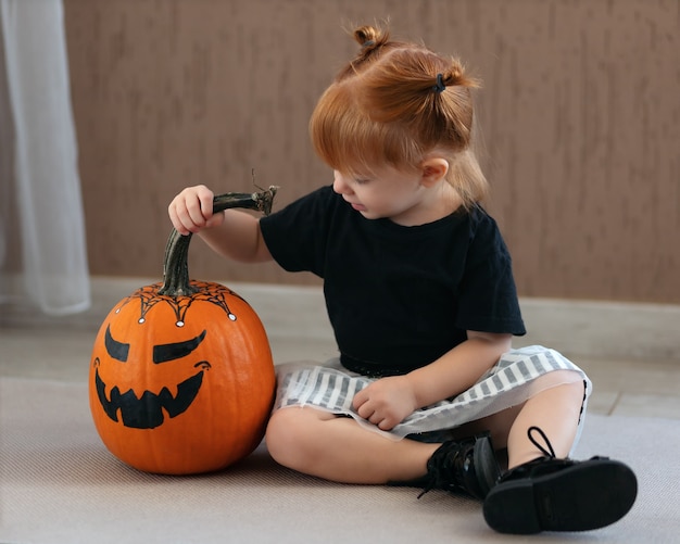 Foto schattig klein meisje zit en kijkt naar een geschilderde pompoen voor halloween in afwachting van de vakantie